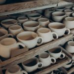 Rows of handmade ceramic cups and bowls on rustic wooden shelves in a pottery studio.
